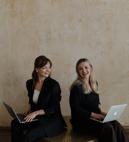 Natalia Balicka i Hanna Juszczak siedzą z laptopami przed ścianą z surowego tynku